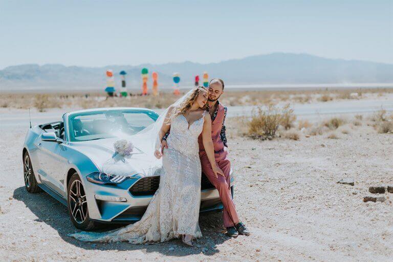 A bride and groom pose on a car in the desert