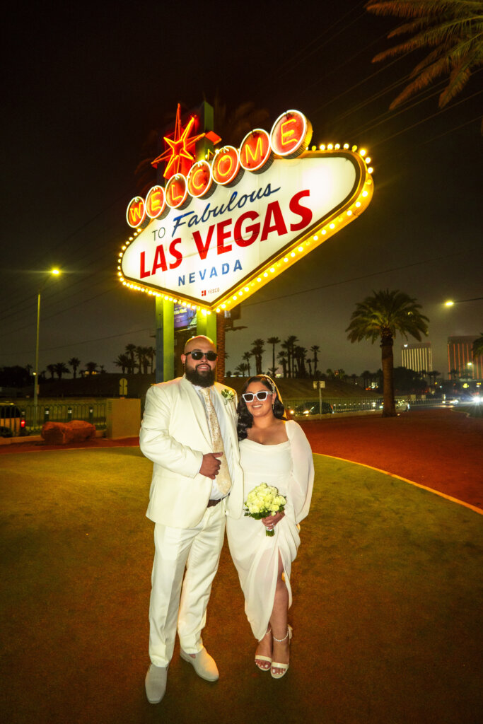 a couple wearing white standing in front of the Fabulous Las Vegas sign