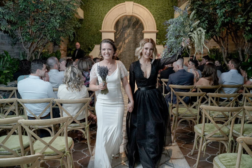 two women walking down a wedding aisle, one in a black dress and one in a white dress