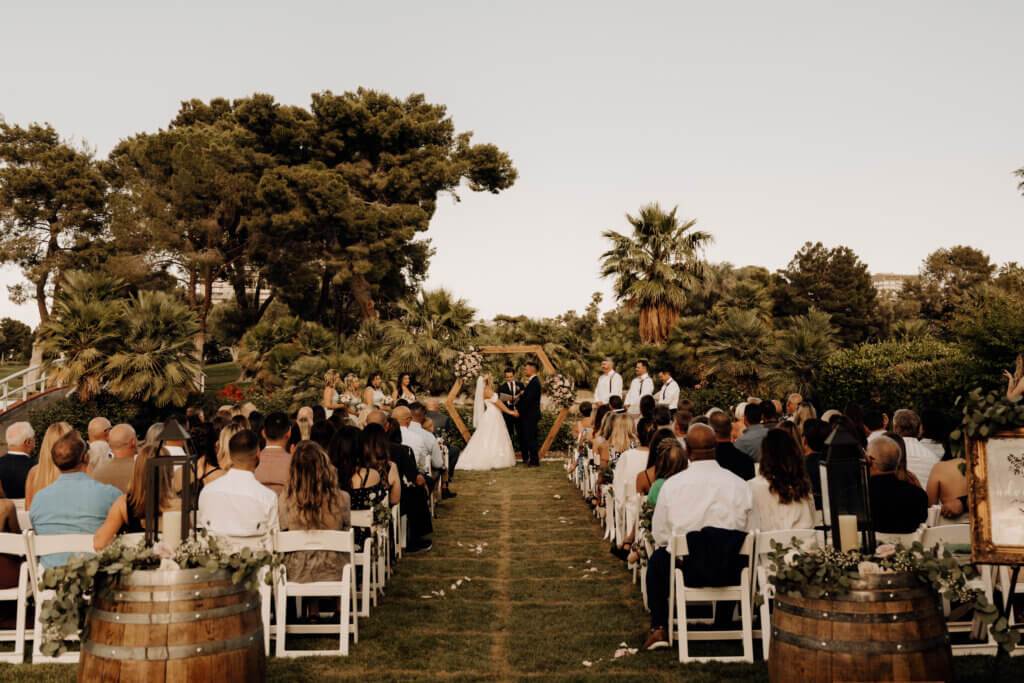 a large outdoor wedding from a back view with a bride and groom holding hands at the altar
